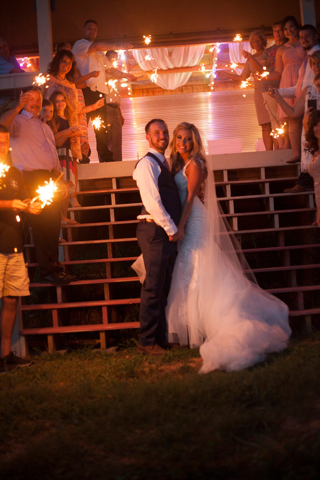A bride and groom pose together at night under warm string lights with wedding guests gathered around them.