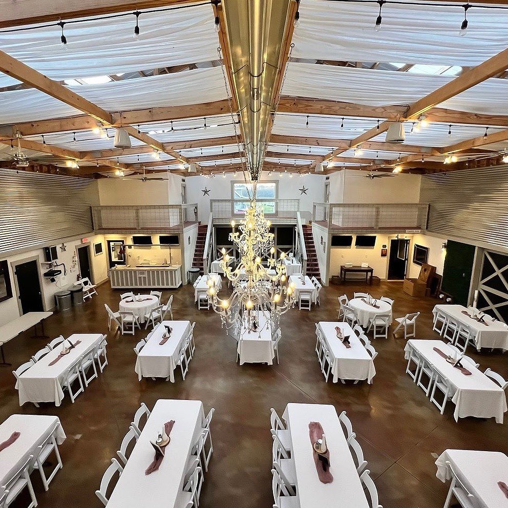 An elegant indoor barn venue with wooden beams, skylights, white linens, and a central chandelier set for a formal event.