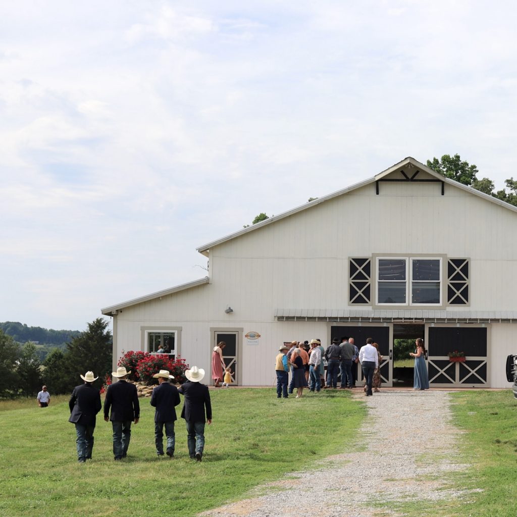 A modern white barn with black X-pattern doors and windows sits on a grassy field with visitors walking toward it.