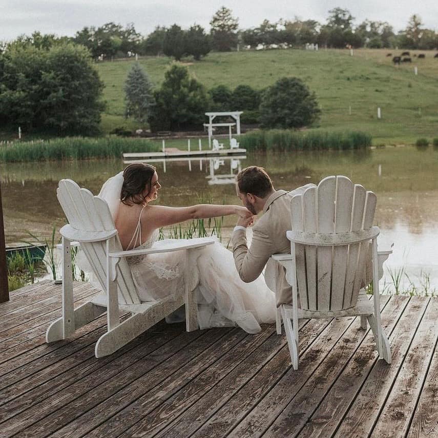 A couple sits in white Adirondack chairs on a wooden dock overlooking a calm pond with pastoral farmland beyond.