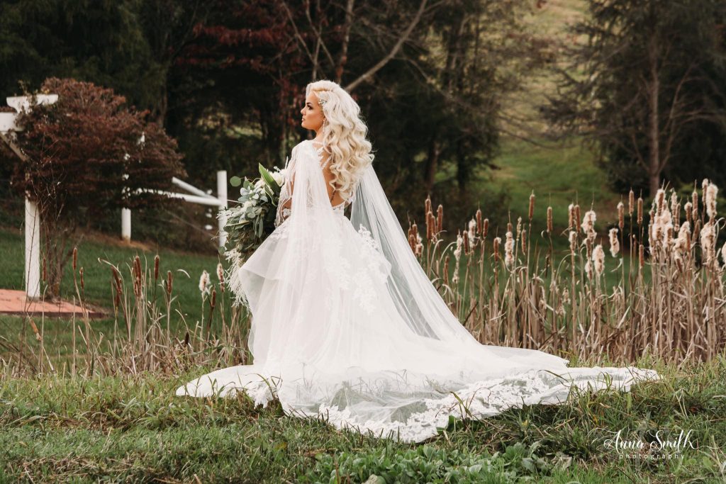 Bride in white gown and veil stands in a grassy field with dried plants and trees in background.
