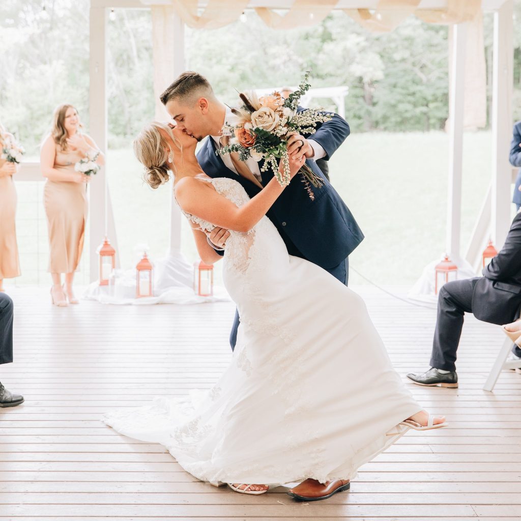 A bride and groom kiss during their first dance on a wooden deck at their wedding celebration.