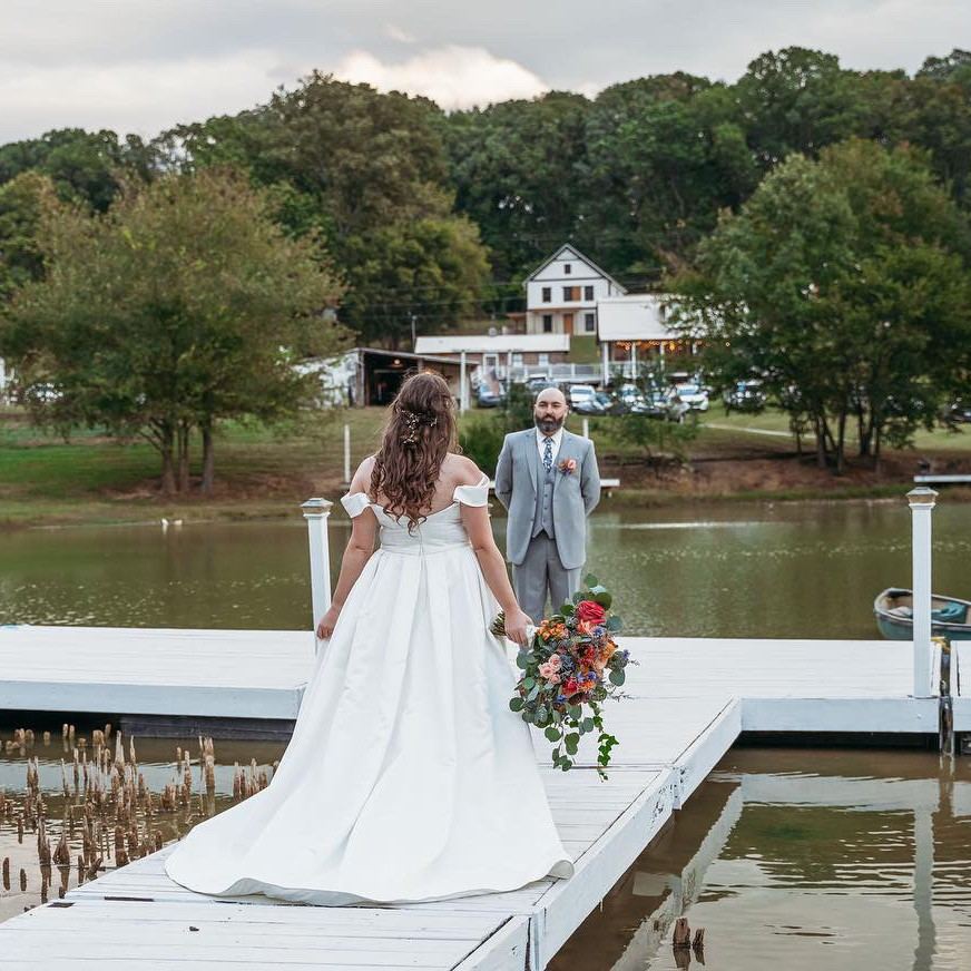A bride and groom stand on a white dock overlooking a calm river with a white farmhouse and trees in the background.
