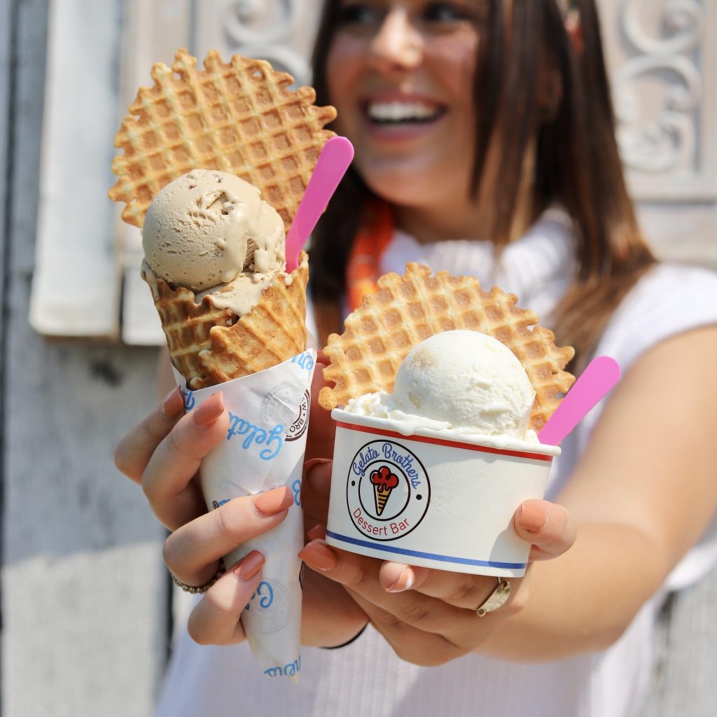 A smiling woman holds two waffle cones with vanilla ice cream scoops in branded cups.