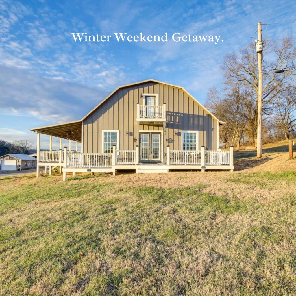 A modern barn-style house with white railings sits on a grassy hillside under a blue sky.