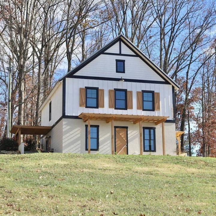A modern farmhouse with white siding and black trim sits on a grassy hillside surrounded by bare trees.