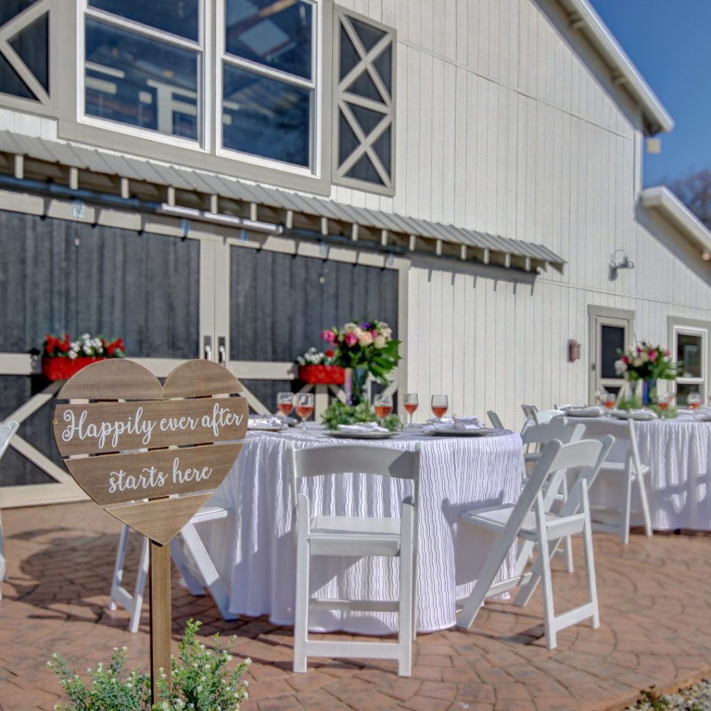Outdoor wedding reception setup with purple-draped tables, white chairs, and floral arrangements on a brick patio beside a white barn.