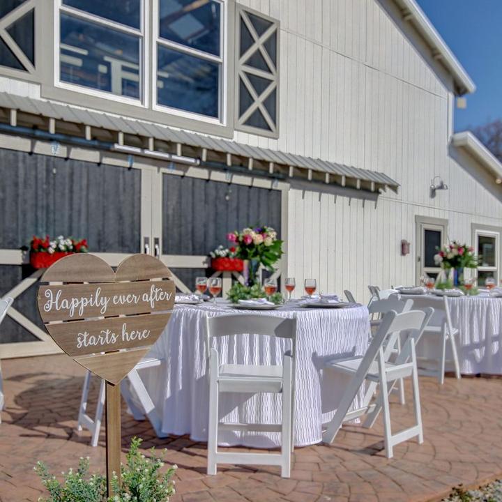 Outdoor wedding reception setup with purple-draped tables, white chairs, and floral arrangements on a brick patio beside a white barn.