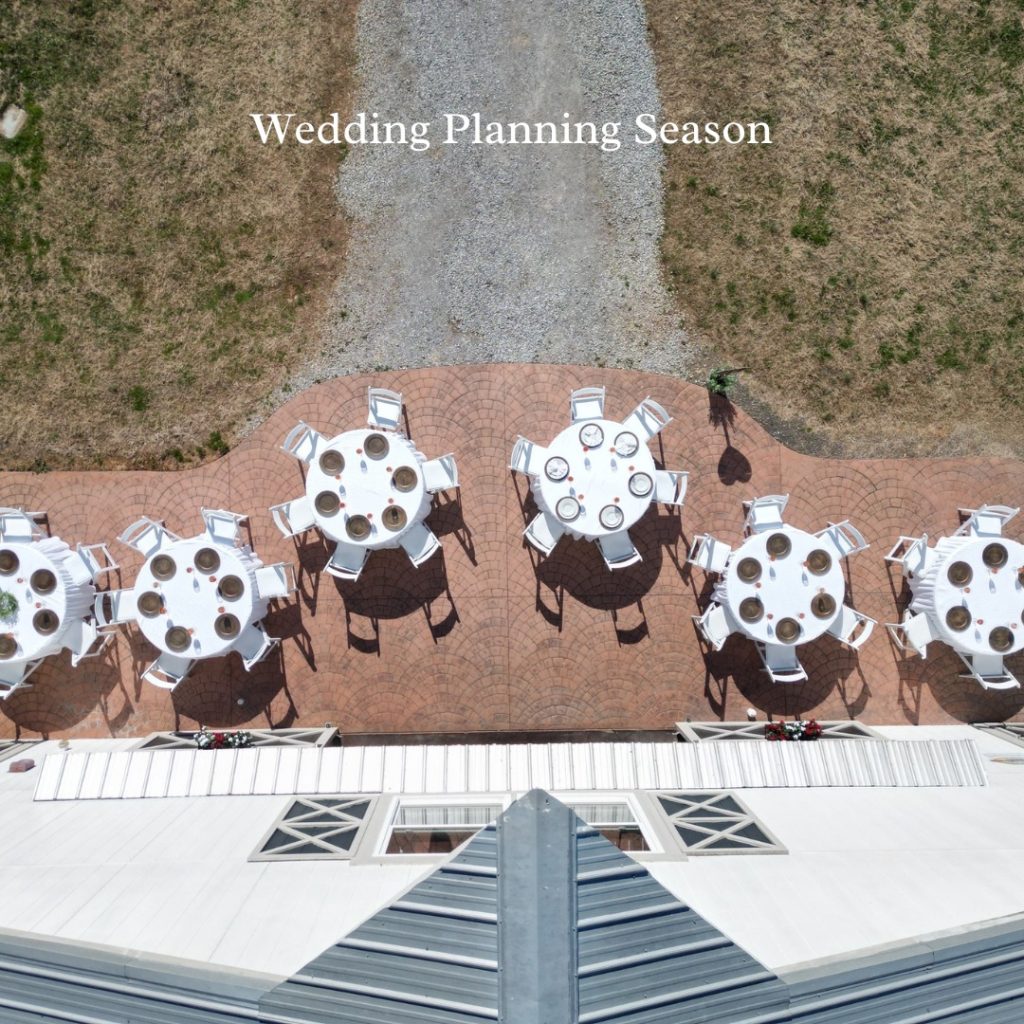 Aerial view of round tables with white linens and brown chairs arranged on a brick patio beside a white building.