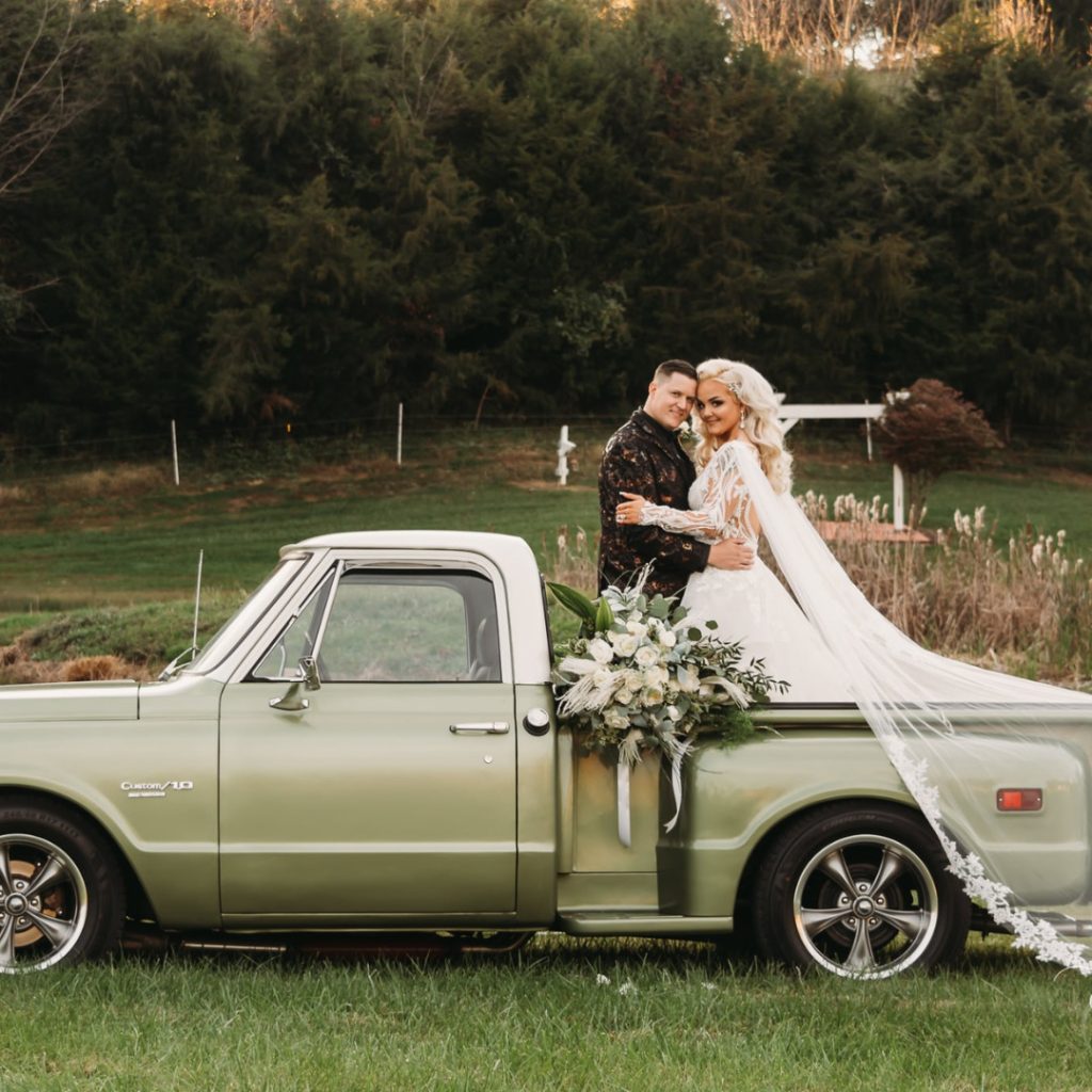 A bride and groom kiss beside a vintage pickup truck decorated with white flowers in a rural setting.