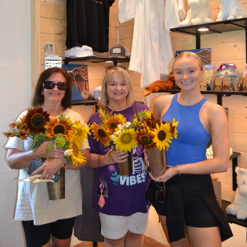 Three women holding sunflower bouquets stand together inside a retail shop space.