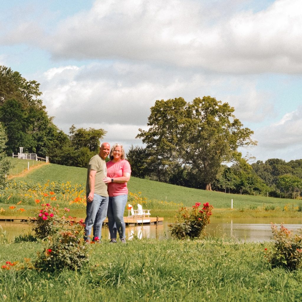 A couple stands together in a pastoral field with wildflowers, trees, and a pond visible in the background.