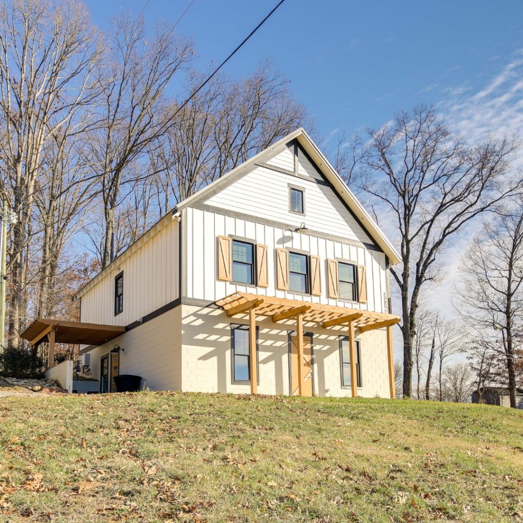 A modern farmhouse with white and cream siding sits on a sloped lawn with bare trees in the background under clear blue sky.