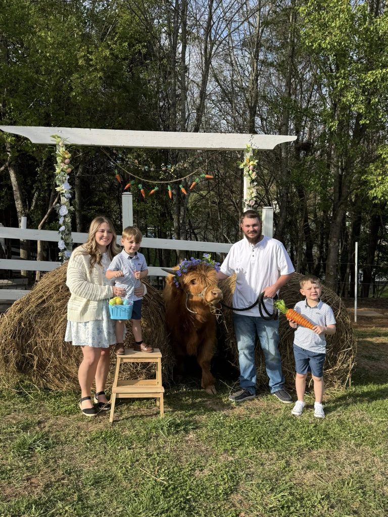 A family of five poses with a brown Highland cow in front of a white wooden structure on a grassy farm.