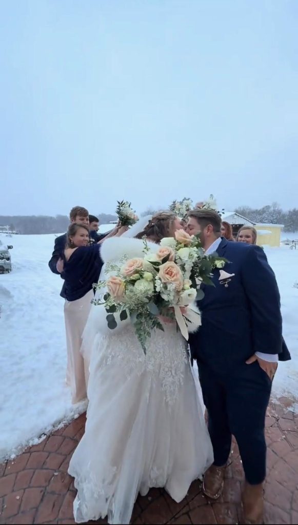 A bride and groom pose outdoors in winter attire with wedding guests and a large floral bouquet on a snowy day.