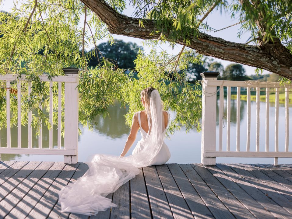 A bride in a long veil sits on the dock over the pond at Cedar Pond Farms, framed by a willow tree.