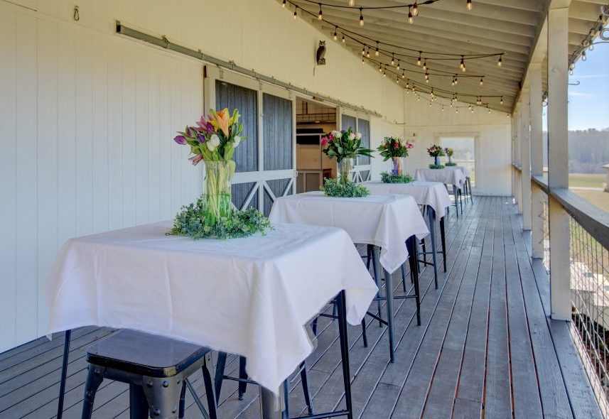 Cocktail tables dressed with spring flowers along the covered porch at Cedar Pond Farms, string lights overhead and the fields beyond.