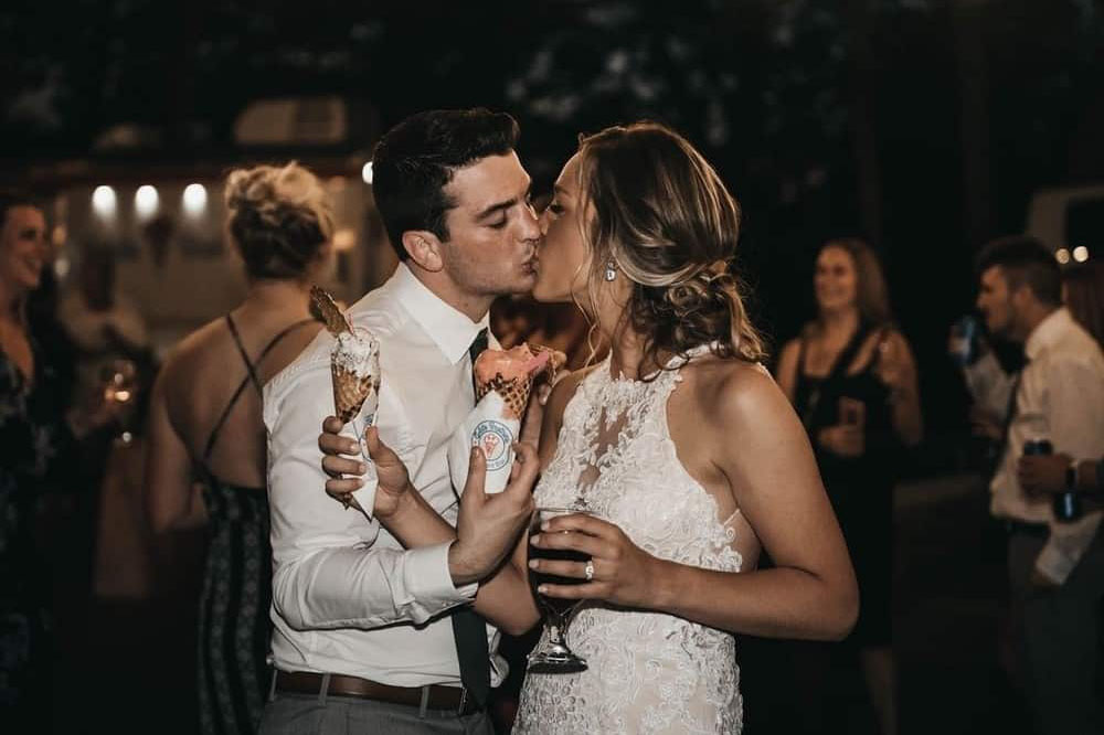 A couple in formal attire kiss while holding champagne glasses at an evening celebration surrounded by guests.