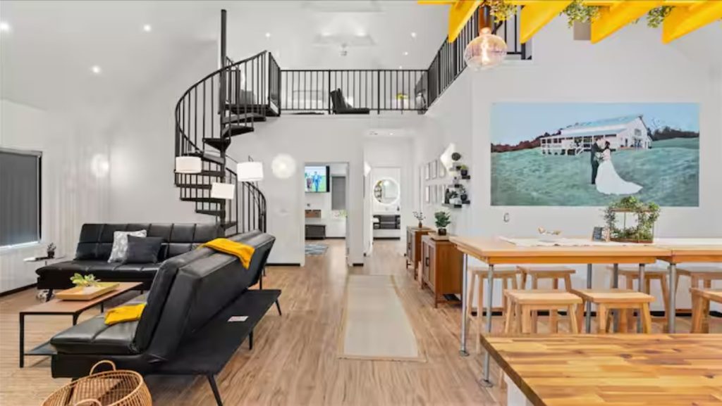 Modern loft interior with spiral staircase, black furniture, wooden dining table, and bride portrait on wall.
