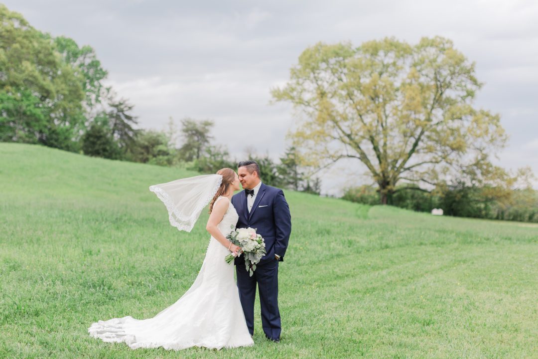 A bride and groom stand together in an open pasture at Cedar Pond Farms, her veil catching the wind.