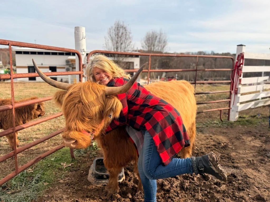 A woman in a red plaid shirt hugs a brown Highland cow in a rural farm pen.