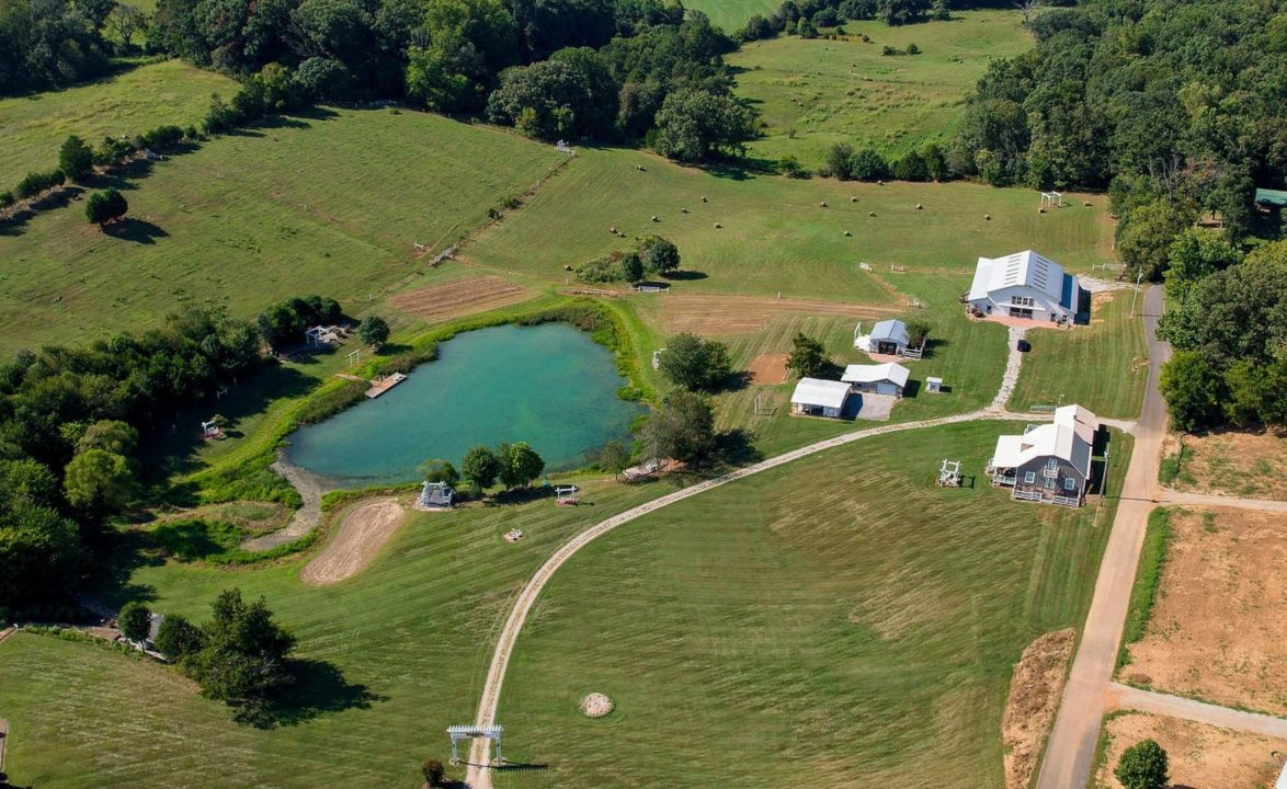 An aerial view of Cedar Pond Farms — the pond, the pastures and the white barns — the Tennessee countryside a hot air balloon rises over at sunrise.