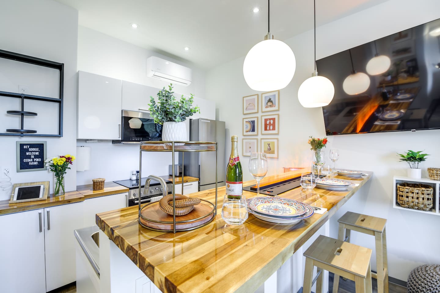Pull up a stool at the butcher-block breakfast bar in this barndominium suite kitchen at the fluffy cow farm — plates set, fresh flowers out and a Cedar Pond Farms welcome board on the counter.